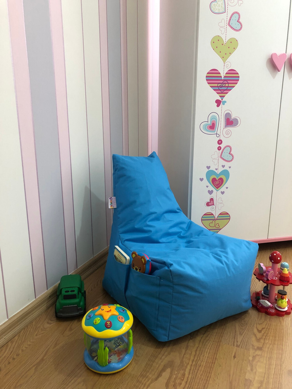 Tirkizna lazy bag vreća s naslonom i bočnim džepovima u dječjoj sobi; pored igračaka i ormara s motivima srca, na drvenom podu – udobno mjesto za čitanje i igru.