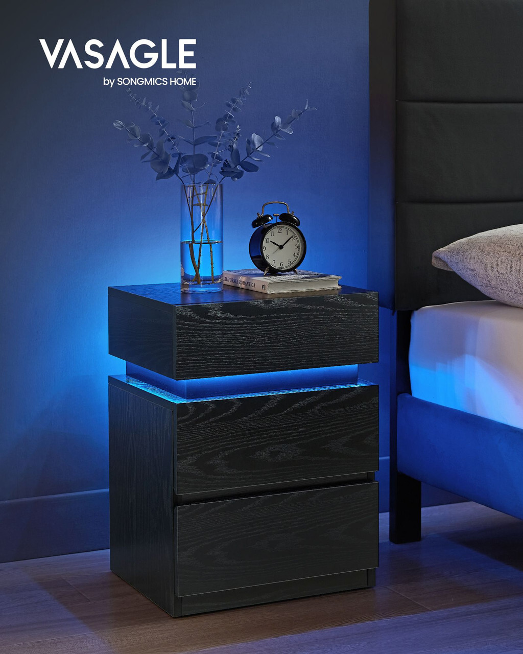 VASAGLE crni noćni ormarić s LED rasvjetom i ladicama uz krevet Crni VASAGLE noćni ormarić s LED osvjetljenjem i dvije ladice, postavljen uz moderan tapecirani krevet.