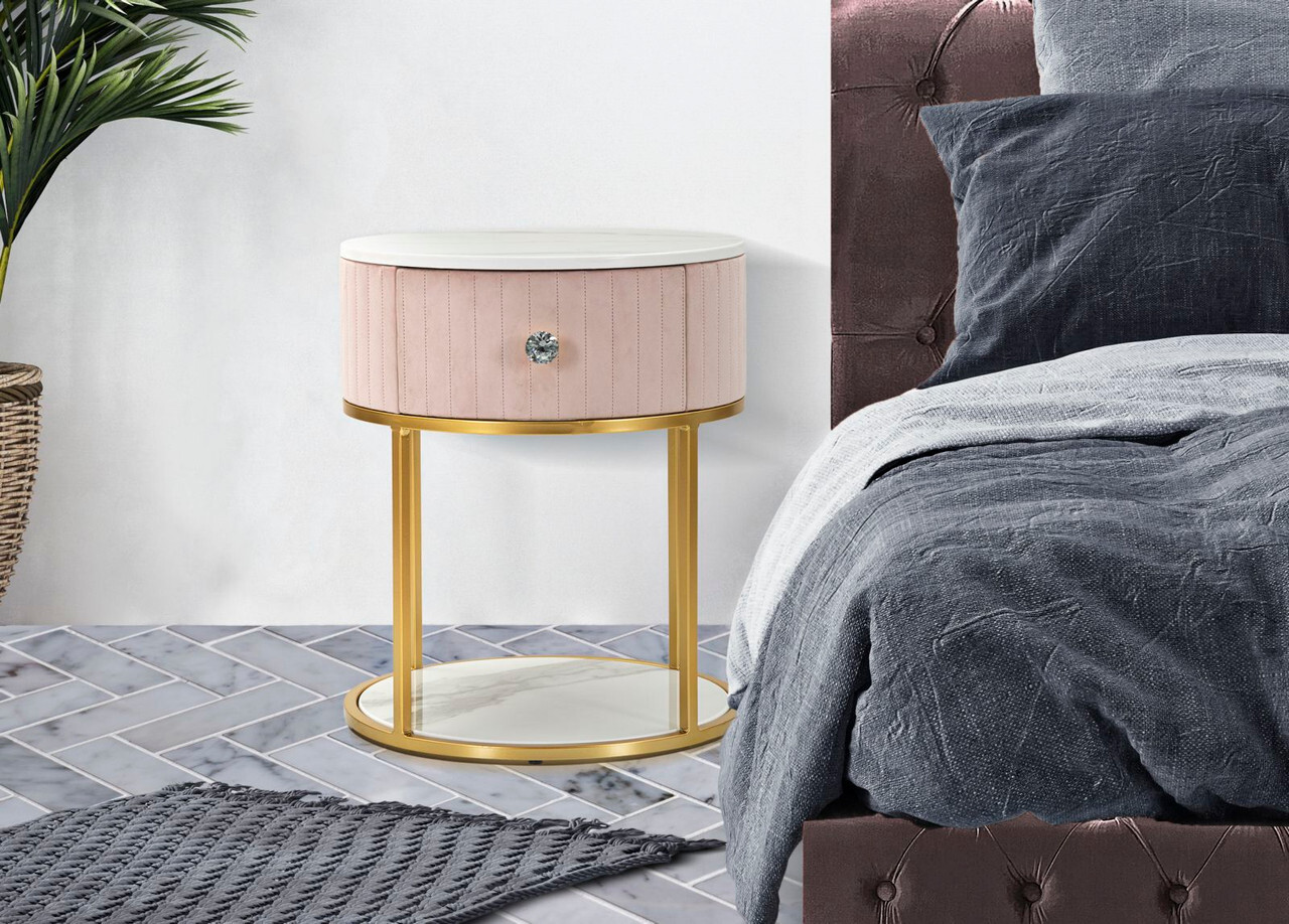 Noćni ormarić Montpellier roza sa zlatnim nogama i ladicom Elegantni roza noćni ormarić Montpellier sa zlatnim metalnim okvirom i ladicom, postavljen uz tapecirani krevet u modernoj spavaćoj sobi.