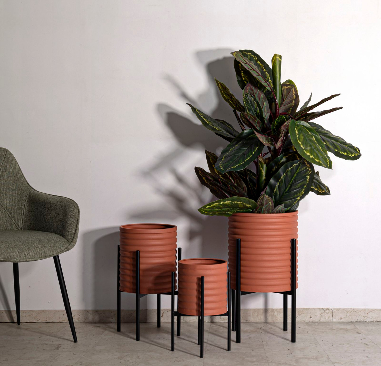 Set od tri terracotta saksije na crnim metalnim stalkima za biljke uz fotelju, idealno rješenje za moderan dnevni boravak i elegantan prikaz kućnog cvijeća.